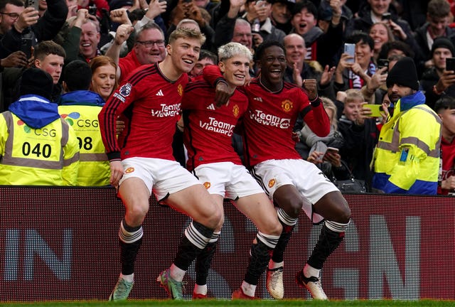 Alejandro Garnacho celebrates after scoring against West Ham, sitting on an advertising hoarding flanked by Rasmus Hojlund, left, and Kobbie Mainoo, right