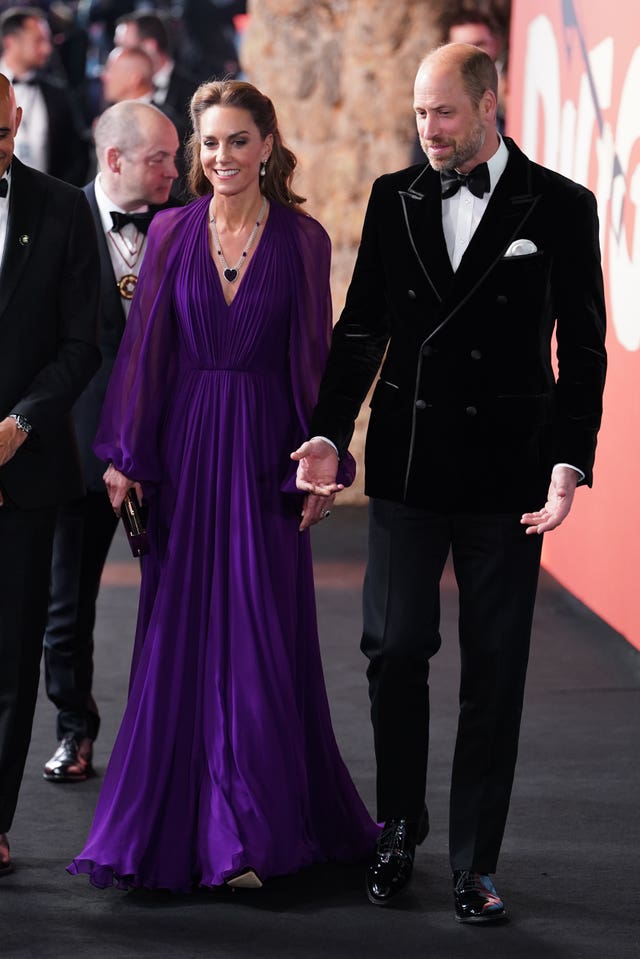 The Princess and Prince of Wales holding hands. She is wearing a long sleeved purple gown while he wears a black suit with bow tie