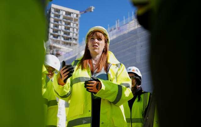 Angela Rayner wearing yellow high-vis jacket and a white hard hat and orange gloves stands in front of a building site with a crane overhead. 