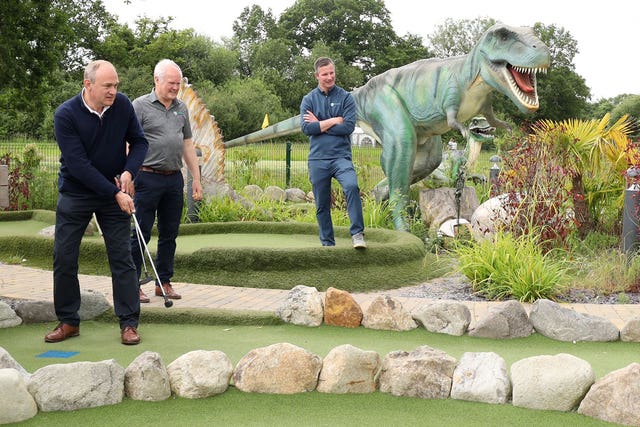 Sir Ed Davey swings his golf club and hits a ball in front of a large dinosaur figure