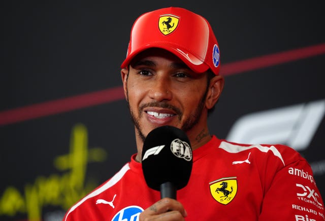 Lewis Hamilton smiling while wearing a red Ferrari jacket and cap holding a microphone
