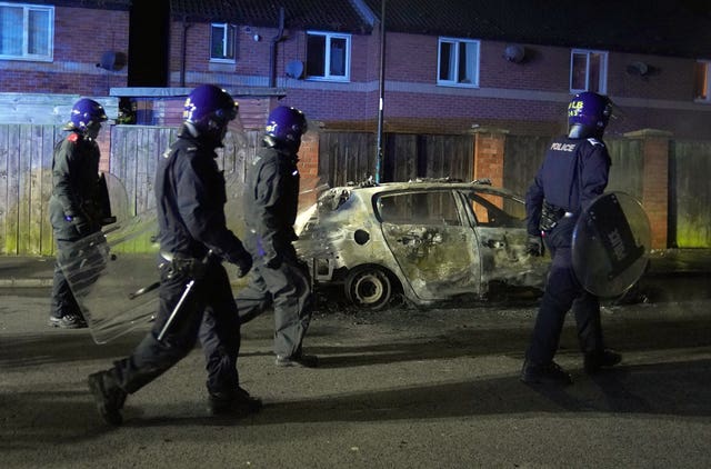 Police officers in riot gear walk past a burnt-out car on a residential street during rioting in Hartlepool.