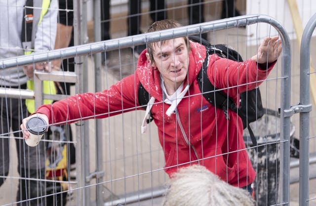 A man in a red hoodie with his arms outstretched behind a metal fence, clutching a coffee cup in his right hand