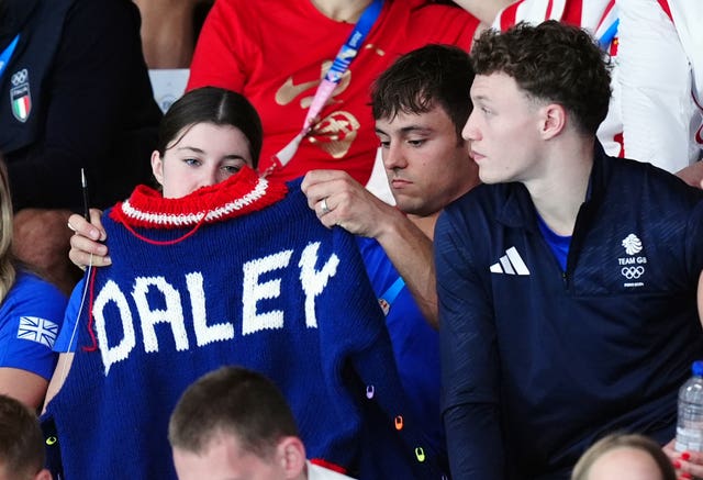 Tom Daley holds up a blue, red and white jumper with his name in white letters 