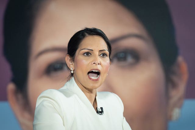 Dame Priti Patel speaking in front of a large picture of herself