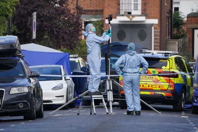 Officers in forensic suits in front of a police car in the middle of a residential street.