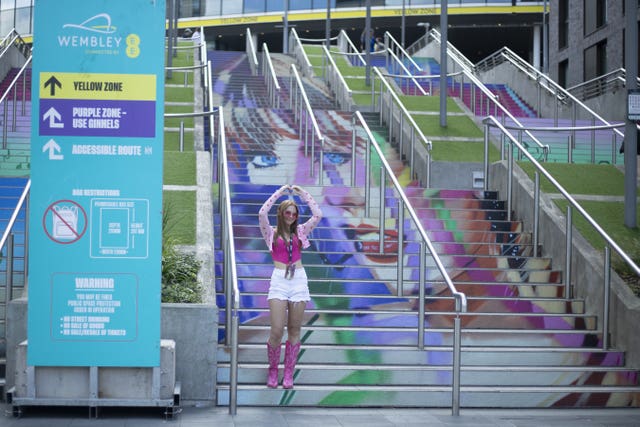 A Taylor Swift fan wearing a pink top and boots poses with her hands above her head forming a heart shape on the steps at Wembley, which feature an image of the singer