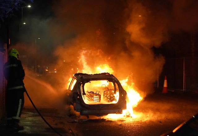 The wreckage of a police car burns as a firefighter sprays water from a hose at it