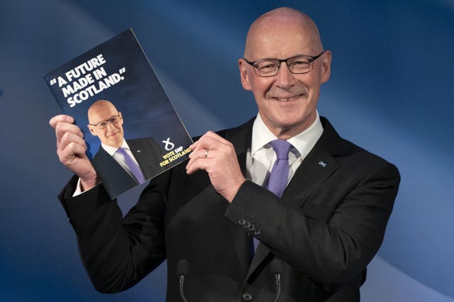 John Swinney smiling while holding up a copy of the SNP manifesto