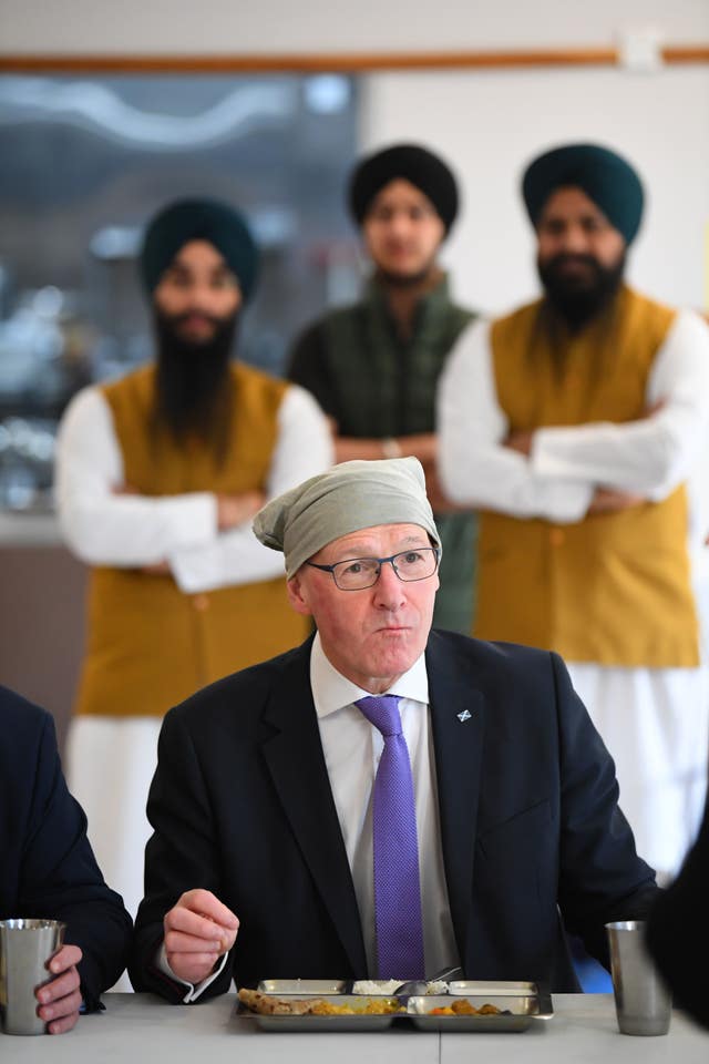 SNP leader John Swinney sits eating curry with his head covered while visiting a Sikh Gurdwara while behind him stand three men in turbans 