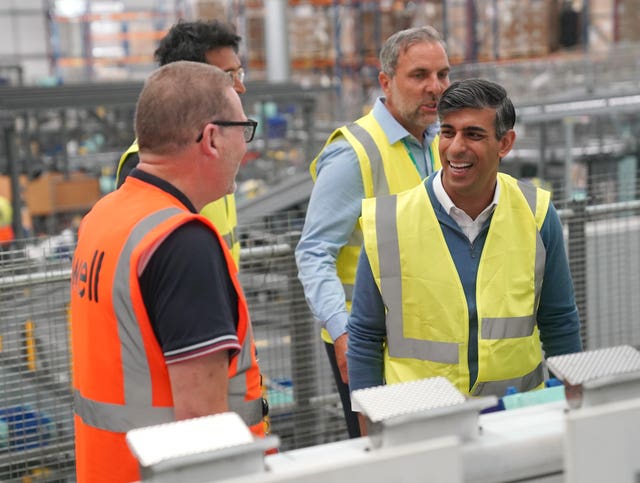 Rishi Sunak in a yellow hi-vis vest laughs in conversation with a man, wearing glasses and an orange hi-vis vest
