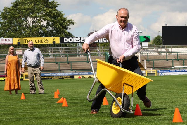 Sir Ed Davey negotiates an obstacle course made out of cones pushing a bright yellow wheelbarrow at speed
