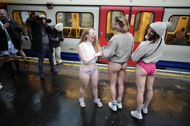 Three women dressed without trousers stand on a tube platform