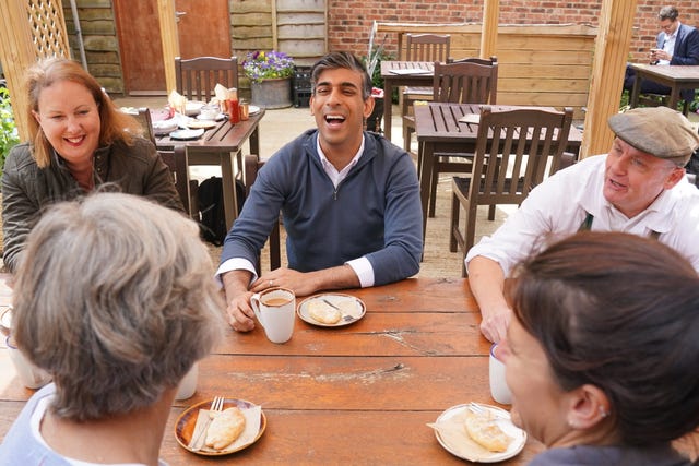 Rishi Sunak seated at a wooden table with four other people with a mug of tea and a plate of food in front of him