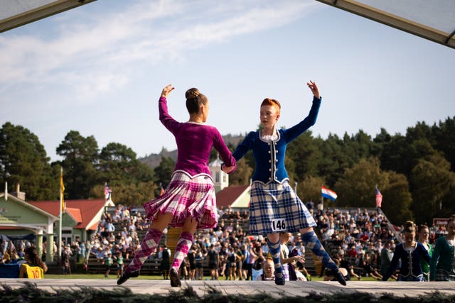 Highland dancers hold their left arms in the air while their right arms are linked as they perform on stage in front of a crowd