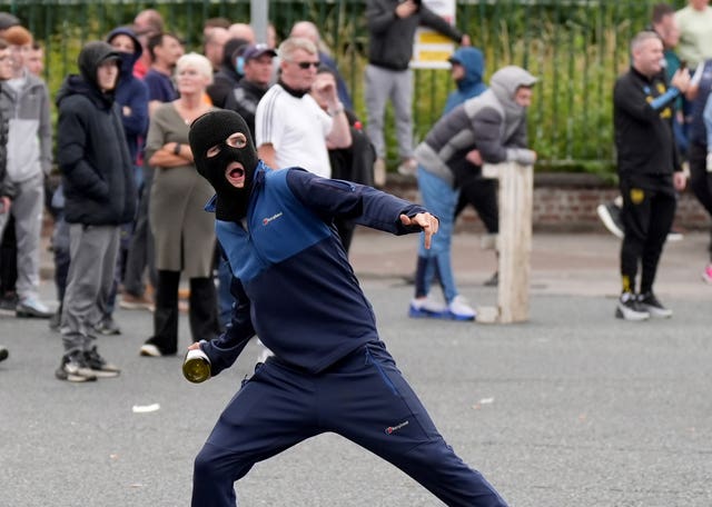 A youngster in a mask prepares to hurl a bottle, while a crowd stands behind him