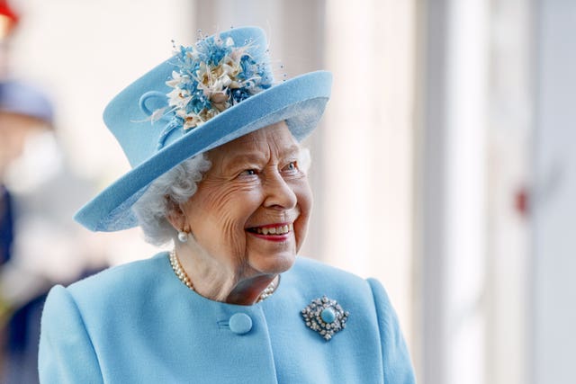 The Queen looks to the right and smiles as she wears a hat decorated with flowers and a diamond jewelled brooch