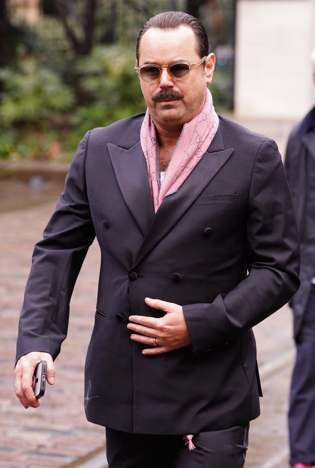 Danny Dyer walking outside, wearing a suit with pink scarf and sunglasses