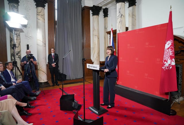 Chancellor Rachel Reeves stands at a lectern in front of a red backdrop featuring the HM Treasury logo for a speech to ministers, business chiefs and reporters at the Treasury 