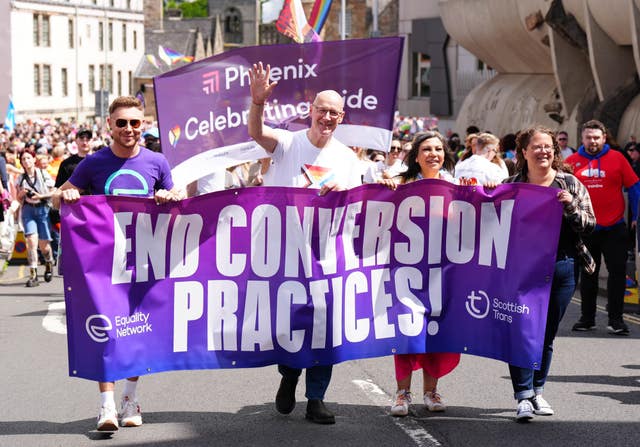 John Swinney wearing a Pride T-shirt holds a banner, with two people, which says End Conversion Practices