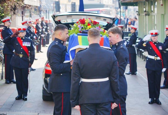 Marines in uniform lift a coffin onto their shoulders. The coffin is draped with a blue flag and has bright flowers on top. Either side of the hearse, men stand in salute