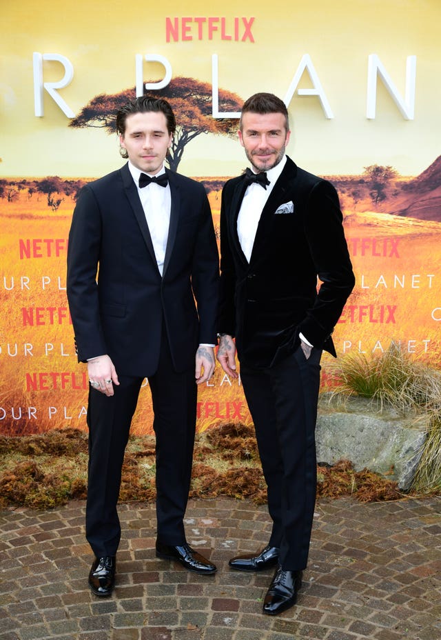 Sir David Beckham with son Brooklyn smiling at a red carpet event, with both dressed in black suits, white shirts with black bow ties