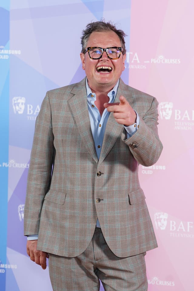 Alan Carr pointing his finger at the camera while laughing and wearing a grey checked suit at the 2026 Baftas.
