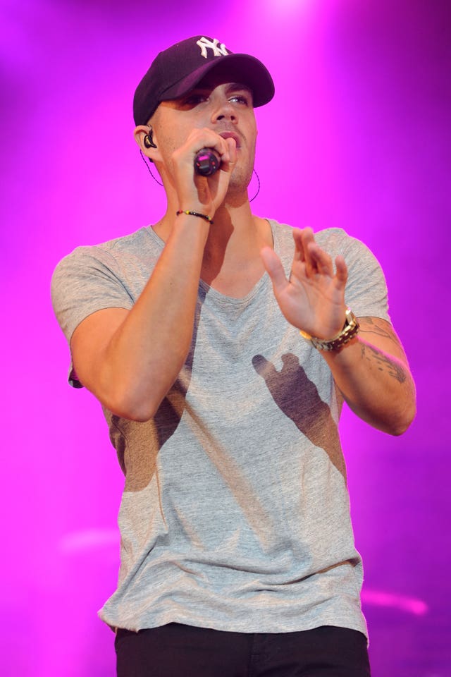 Max George performing on stage, wearing a grey T-shirt and blue baseball cap