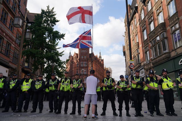 A line of police face a single protester who has their back to the camera and carries a St George's Cross and a Union Flag