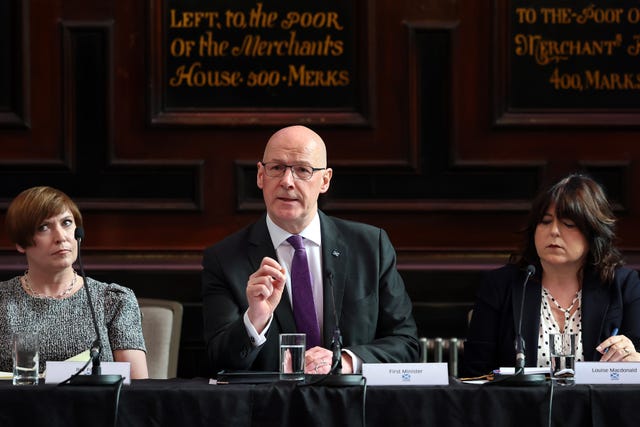 John Swinney gesturing with one hand while speaking from a table, flanked by two women