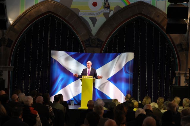 John Swinney speaking to a hall of people from behind a podium in front of a Scottish flag backdrop