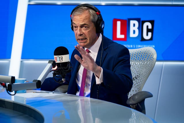 Nigel Farage wearing headphones is sat in front of a microphone. Behind him is a blue background and a logo for LBC radio