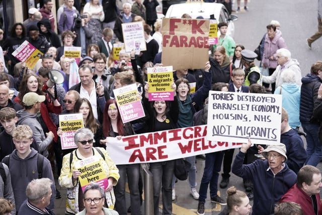 A crowd seen from above holding posters and banners saying refugees and immigrants are welcome, and criticising the Tories and Nigel Farage 