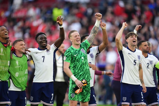 A group of football players in white shirts, the goalkeeper in green is in the centre, they are all smiling with their arms in the air in celebration