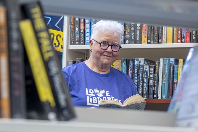 Author Val McDermid wearing a blue t-shirt with a 'love libraries' slogan on it and holding a book, while looking at the camera through an empty space on a shelf of books