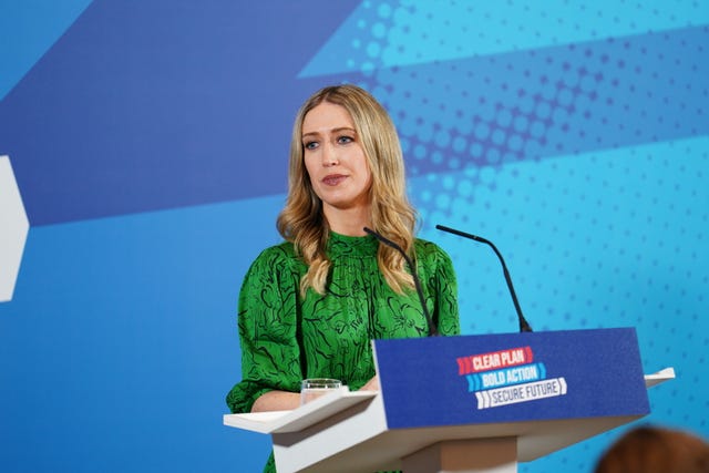 Laura Trott wearing a green dress stands behind a lectern during a Conservative Party election event. There is a blue background and the lectern says "Clear Plan, Bold Action, Secure Future"