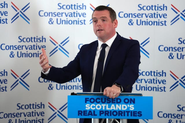 Douglas Ross gestures with his hand while speaking from a lectern featuring Conservative Party signage