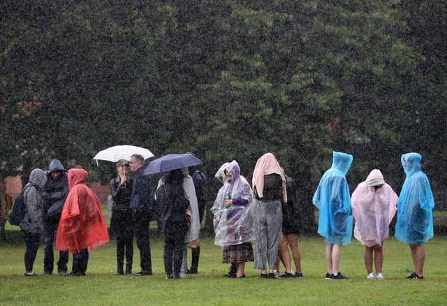 A group of people in ponchos and under umbrellas stand on grass in the rain