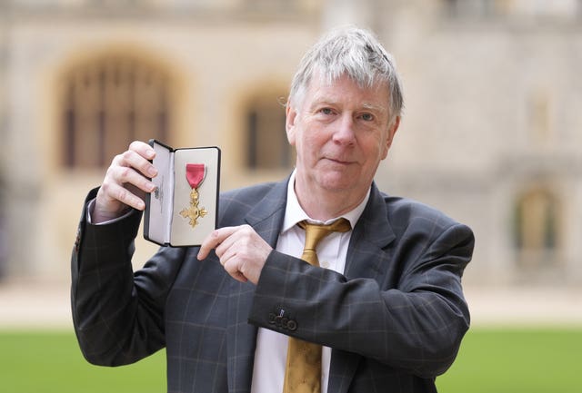 John Whiston posing for a photo while holding up his OBE medal