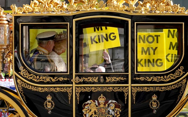 The state coach carrying the King and Queen passes by anti-monarchy protesters waving placards saying 'Not My King'