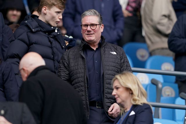 Keir Starmer in the stands of the Etihad Stadium, wearing a puffer jacket, next to a railing. The Manchester City emblem is visible on a seat next to him.