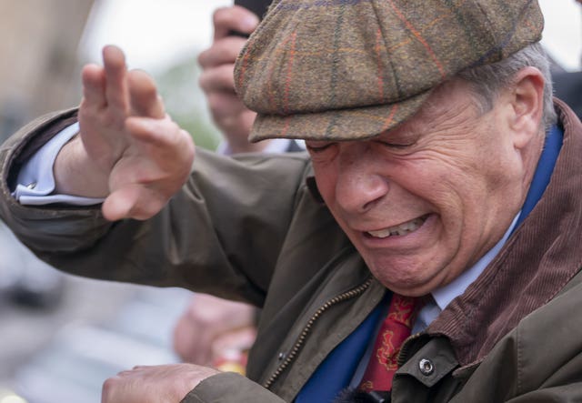 Nigel Farage in a tweedy flat cap flinches with his had over his face as an object is thrown