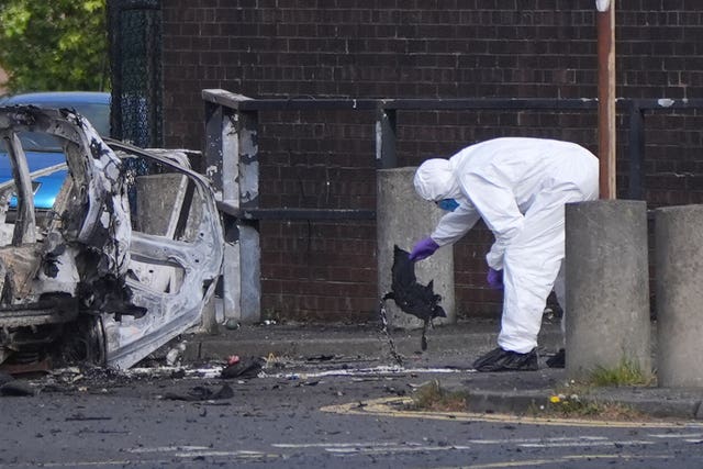 A forensic investigator in a white suit looks at the wreckage of an exploded car