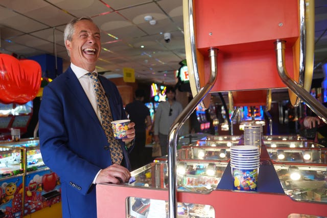 Nigel Farage laughing in a penny arcade while holding a pot of coins over a machine 