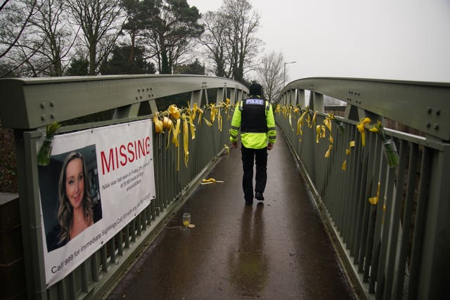 A police officer walks past a missing person appeal poster for Nicola Bulley and yellow ribbons and messages of hope tied to a bridge over the River Wyre in St Michael’s