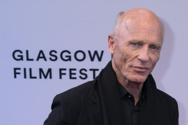 Ed Harris standing in front of a white background with the words Glasgow Film Festival written on it in black writing.