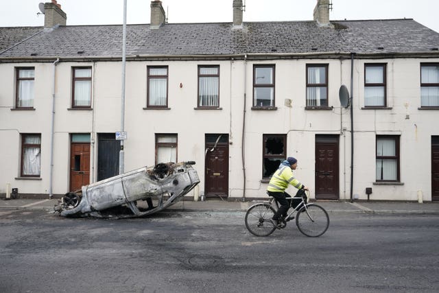 A man cycles past a burnt-out overturned car on the street following a second night of violence in Ballymena