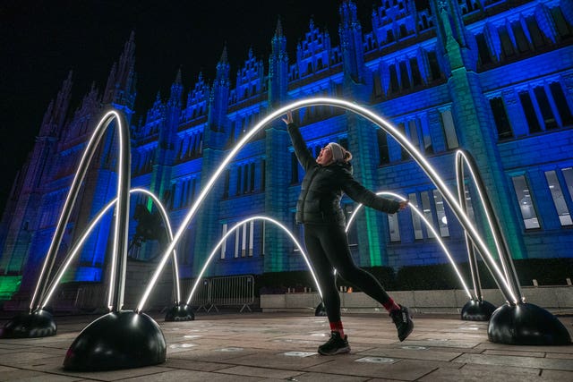 A woman reaches to touch an arc light in front of a building lit in blue