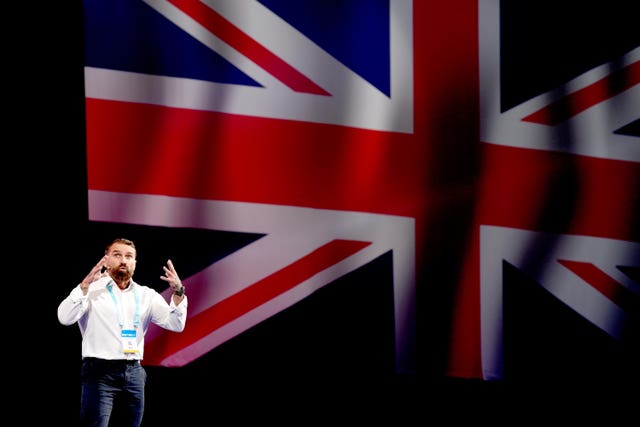 Ant Middleton, a dark haired and dark bearded man, standing on a stage. He is in a white shirt and it contrasts against a huge Union flag