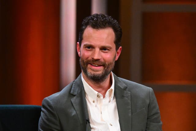 Jamie Dornan smiling wearing a grey suit and white shirt